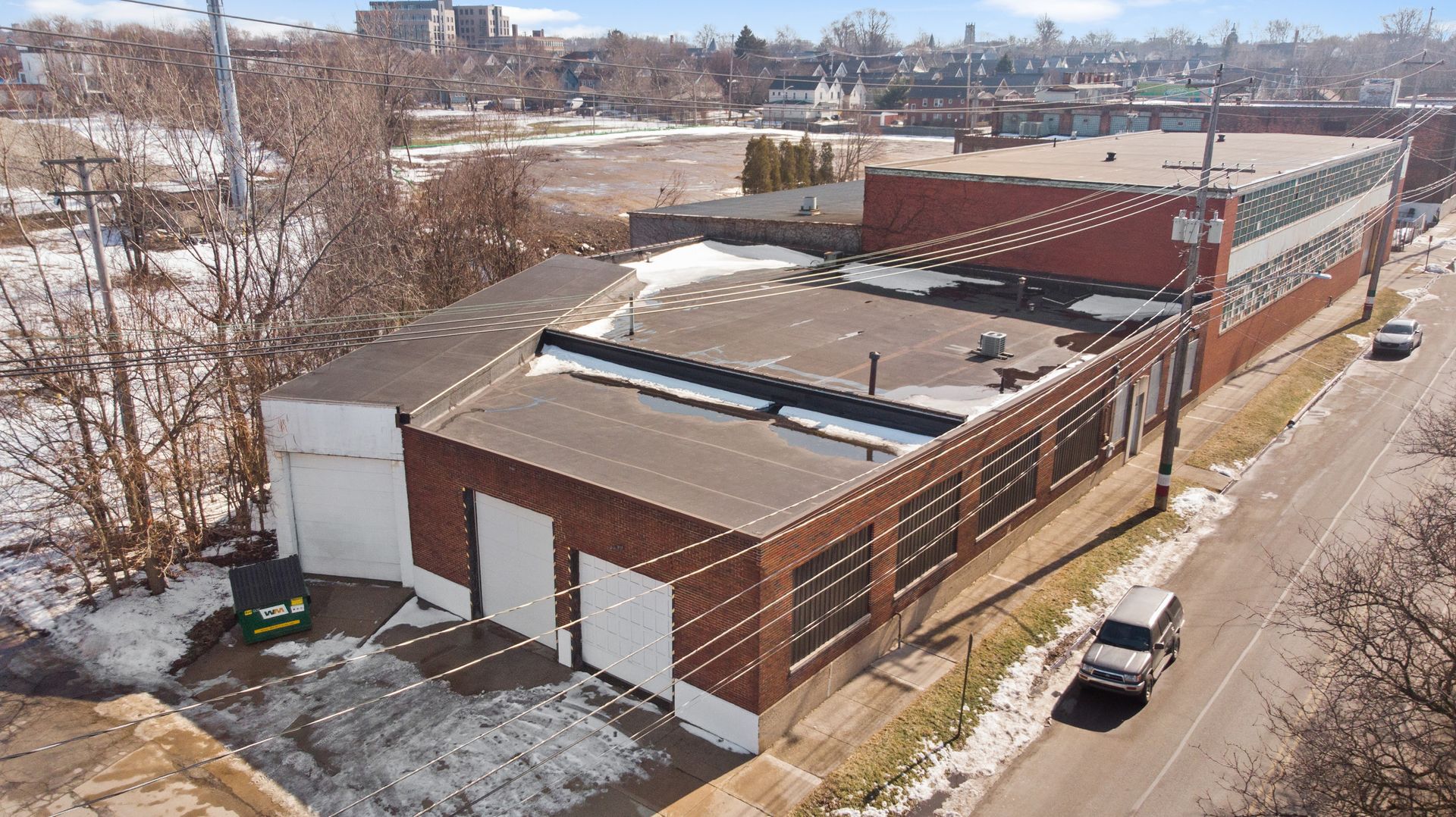 An aerial view of a large brick industrial building with garage doors, flanked by a snow-dusted street and utility poles.