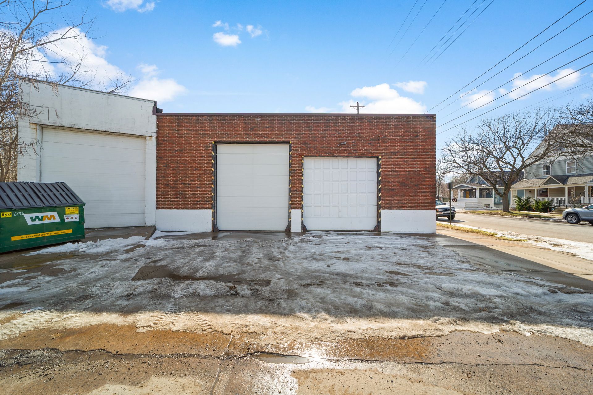 A brick building with three garage doors and a green dumpster on a snowy concrete lot.