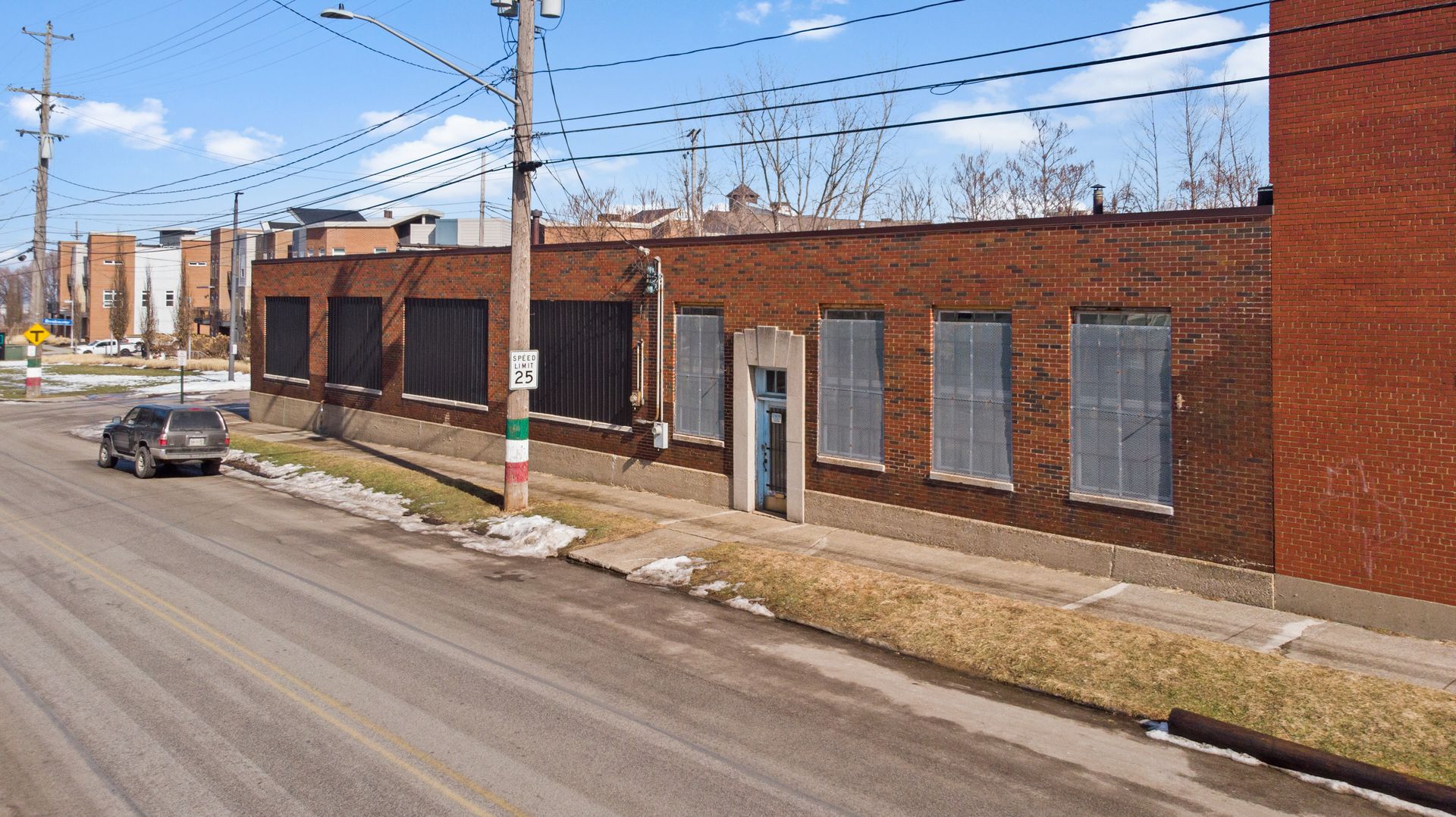 A brick industrial building with barred windows and a speed limit sign on a street corner, with a parked SUV and snow on the ground.
