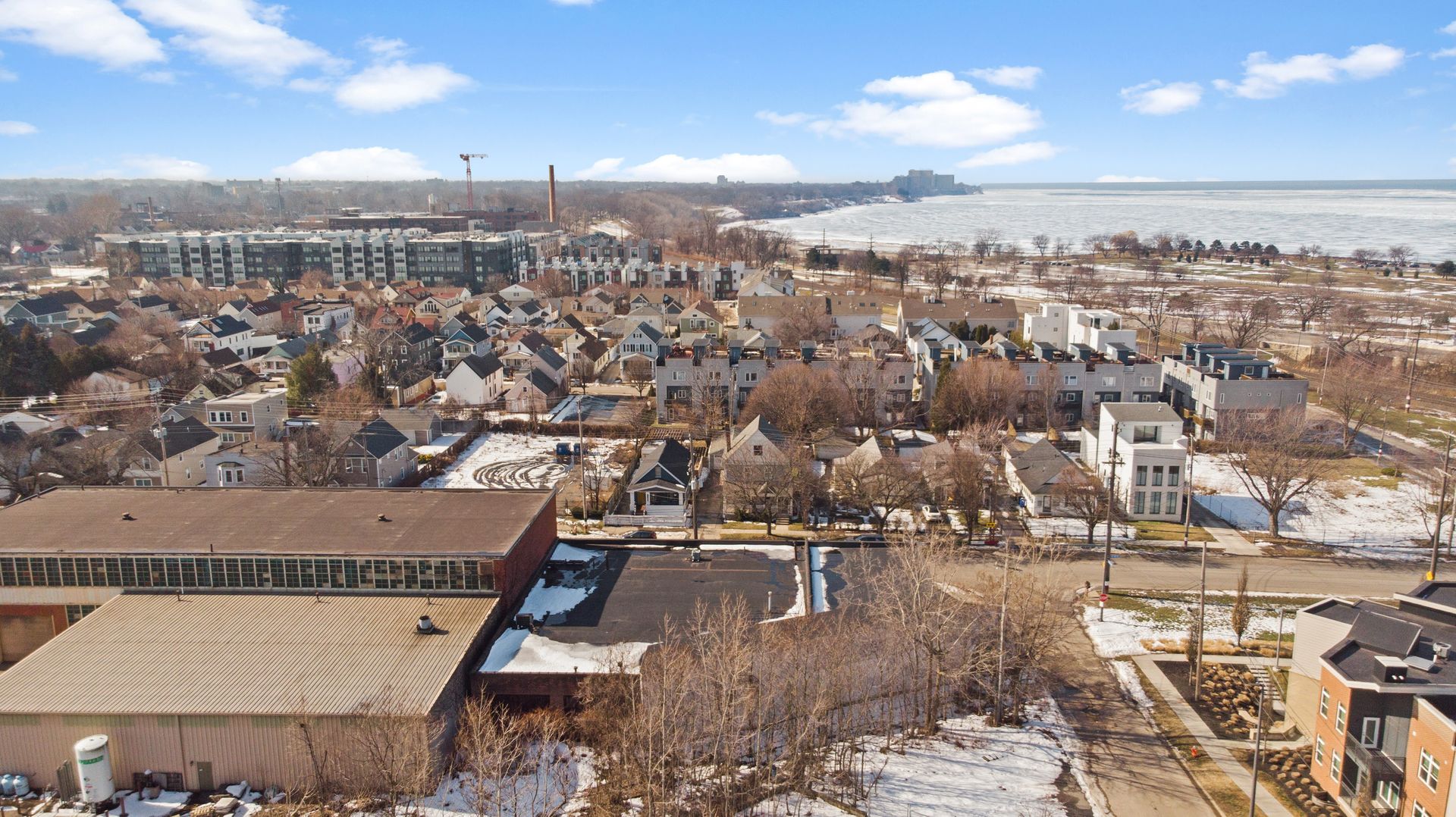 Aerial view of a snowy suburban neighborhood with houses, a large warehouse, and a partially frozen body of water in the background.