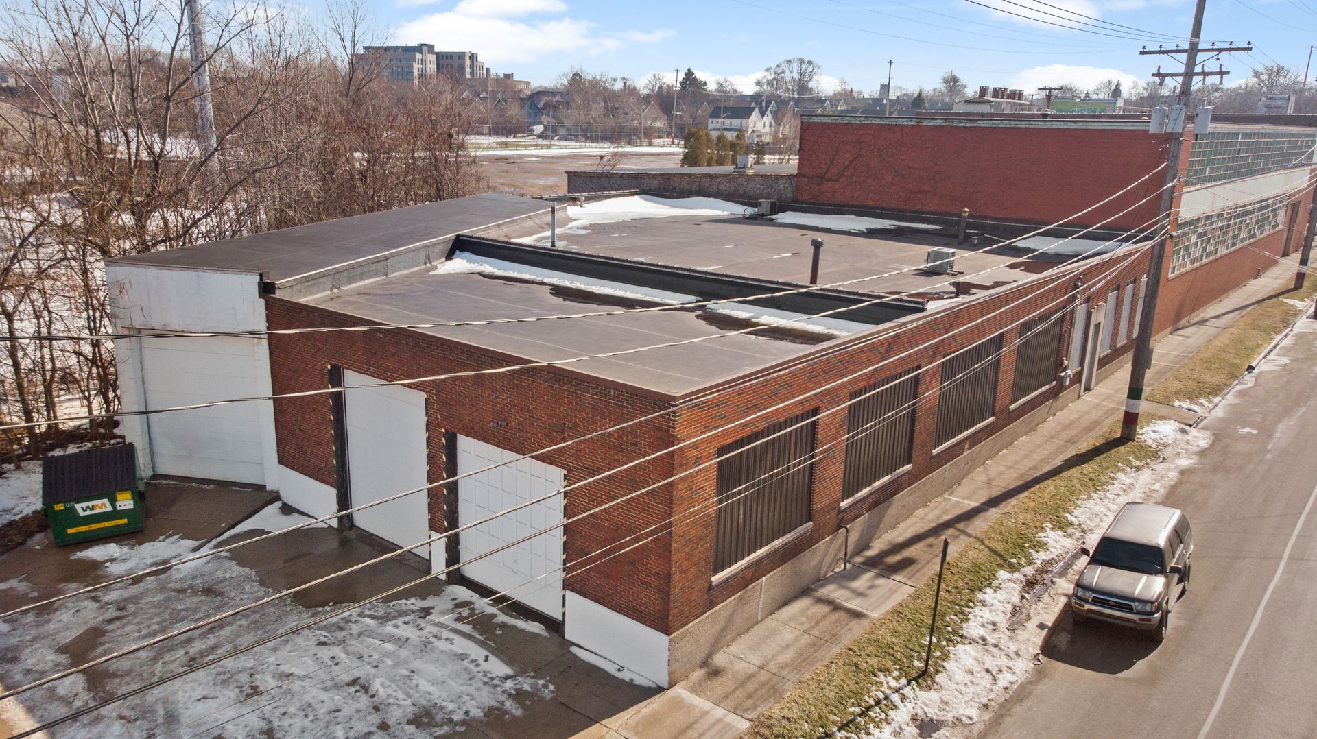 Aerial view of a large industrial brick building with a flat roof and several garage doors, situated alongside a street with parked cars and sparse snow patches.