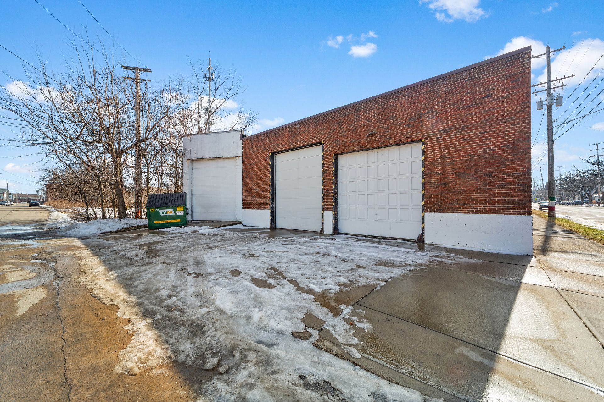 A brick building with three large garage doors and a green dumpster beside a snow-covered driveway.