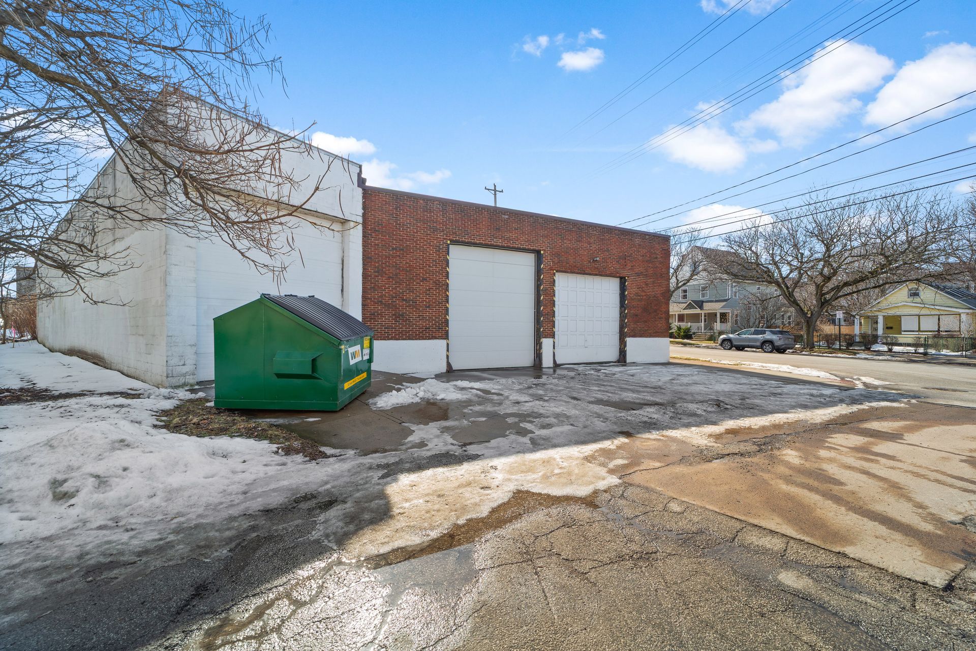 A red brick building with two white garage doors, a green dumpster, and patches of snow on the ground.
