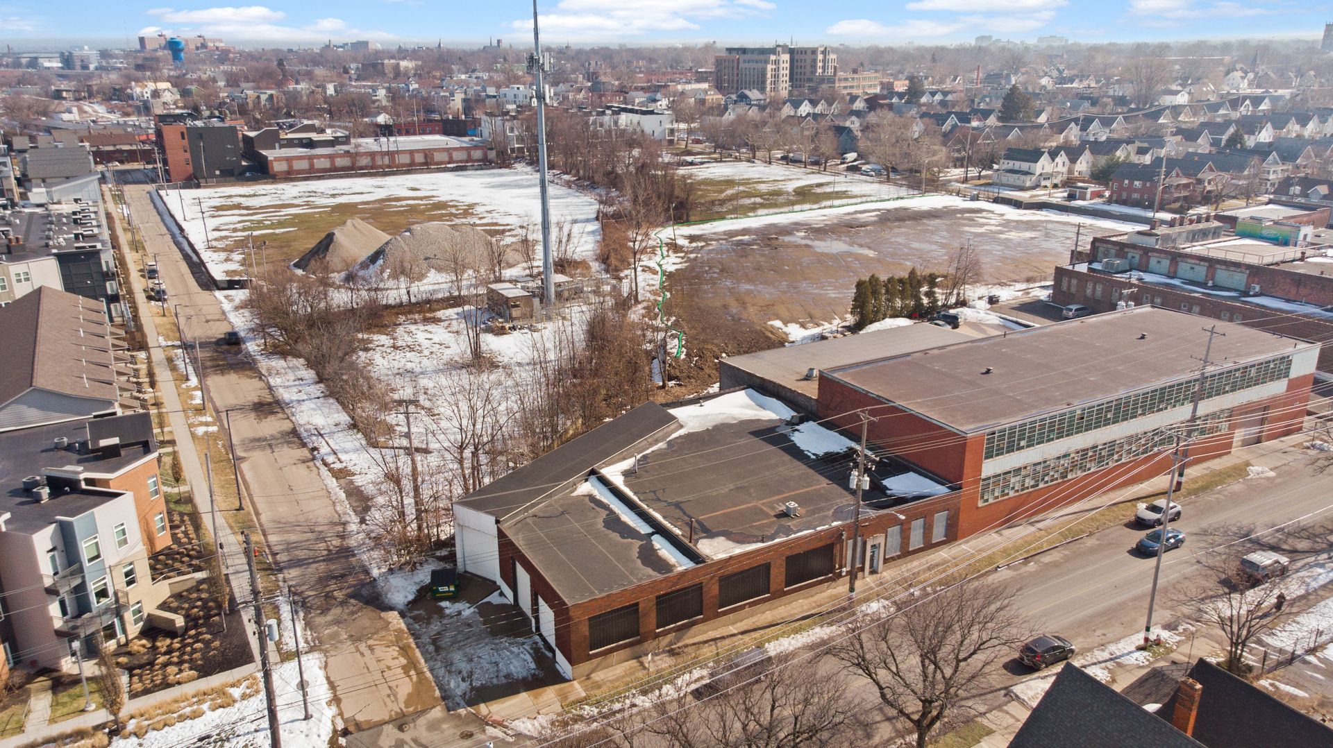 An aerial view of an industrial area with a large brick building, adjacent snowy lots, and nearby residential housing.