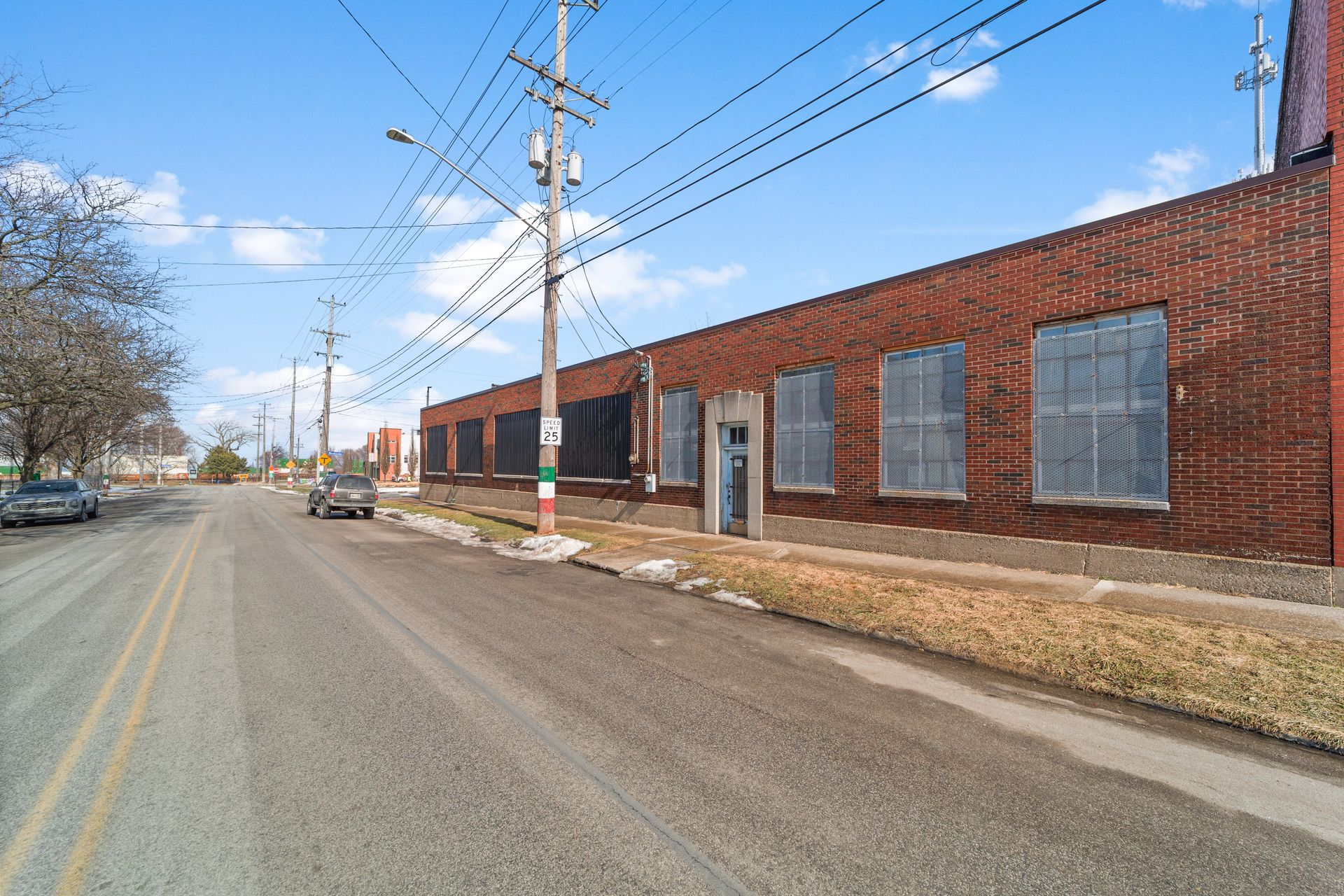 A red brick industrial building lines a street with parked cars and utility poles under a clear blue sky.