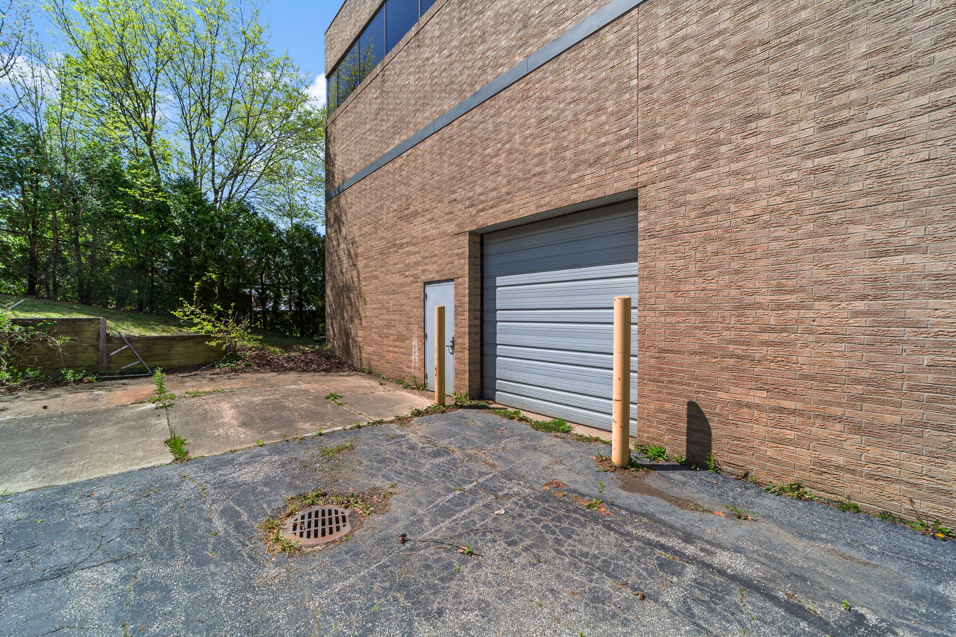 Side view of a brick commercial building with a gray roll-up loading door, a small metal door, yellow bollards, and a cracked paved area beside trees.