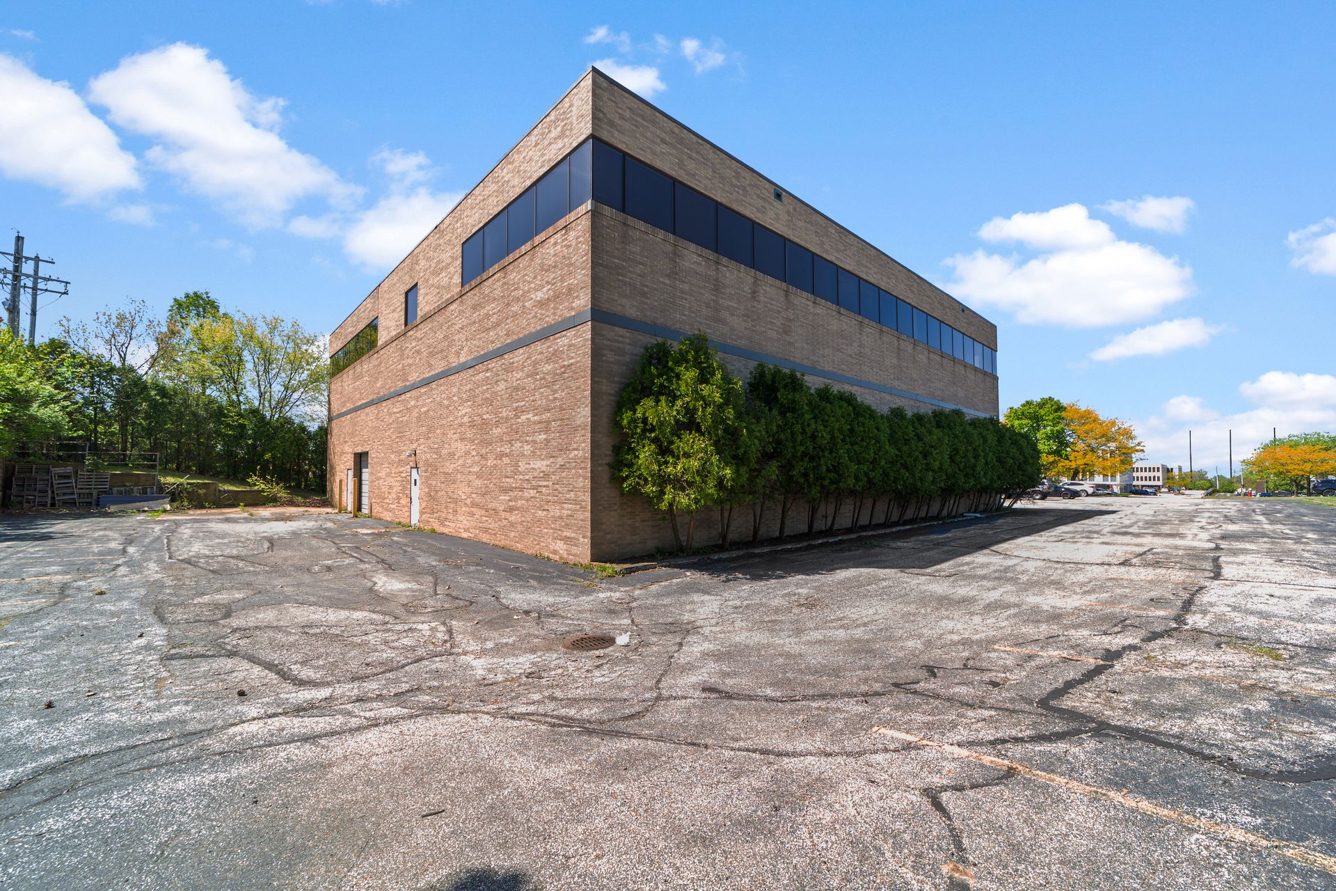 Two-story brick commercial building with dark-tinted upper windows, trees along one side, and a large cracked parking lot under a blue sky.