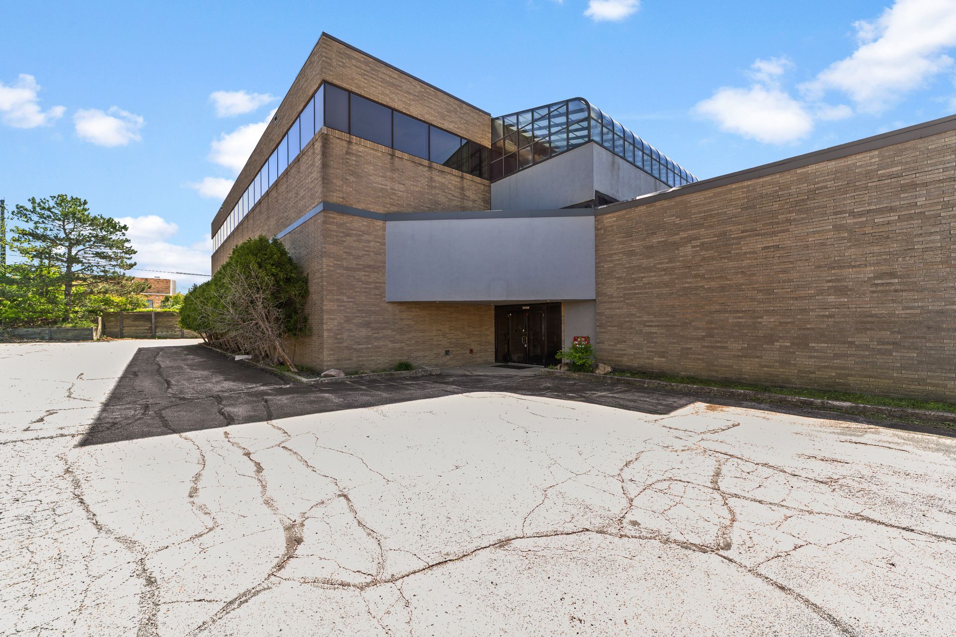 Exterior of a large tan brick commercial building with dark-tinted windows, a glass-roofed upper walkway, and a cracked parking lot in front.