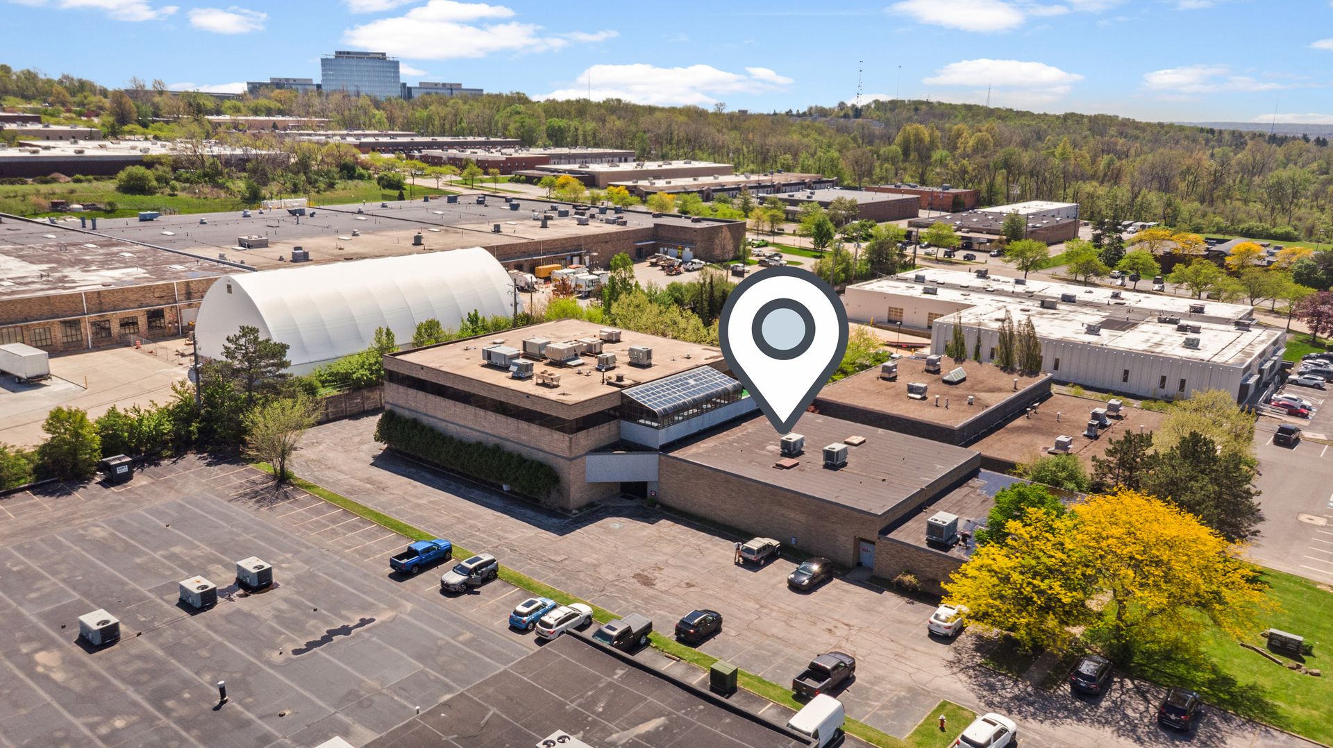 Aerial view of a commercial industrial complex with flat-roof buildings, parking lots, cars, and a large map pin marker over one building.